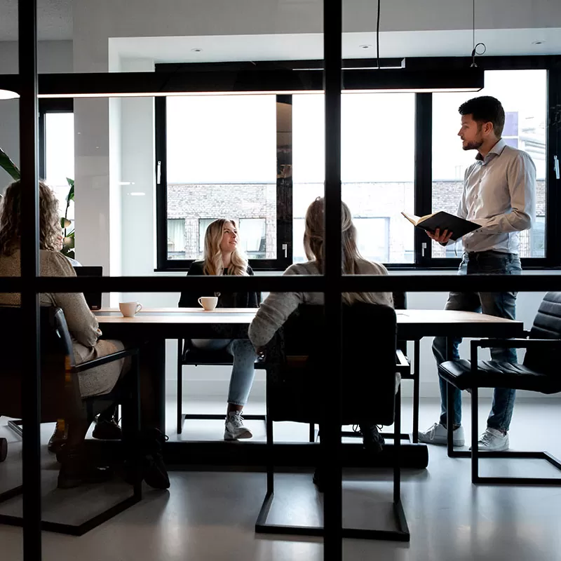 Zakelijke meeting in een vergaderruimte met glazen wanden, waarbij een man een presentatie geeft aan drie collega’s.